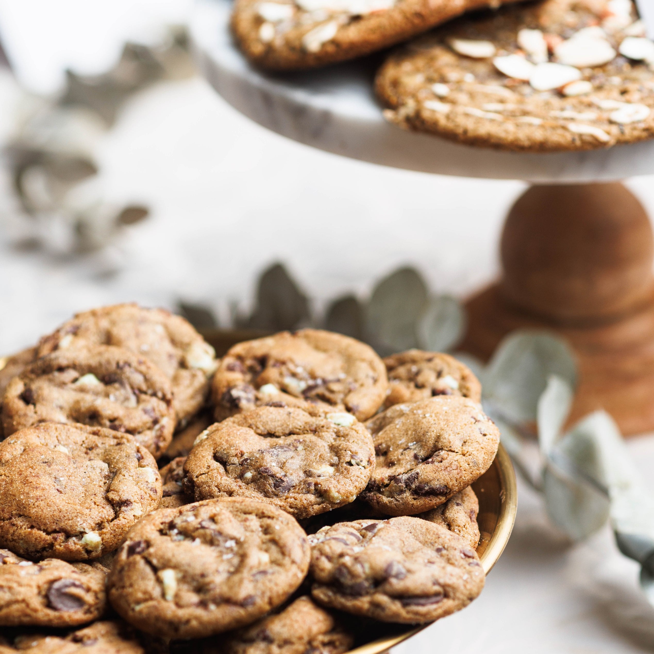 Cake Stand full of Mint Chocolate Chip Cookies with Andes Mints speckled throughout.  A cake stand of Earl Grey Cookies in the background shingled on eachother with Almonds and Fig jam.  The cookies are gooey, soft and chewy, and full of chocolatey goodness. The Andes Mint Chocolate Chip Cookies are perfectly baked, with crinkly tops and sea salt sprinkled all over.  Best Chocolate Chip Cookies, absolutely delicious and the best chocolate chip cookie recipe ever. 